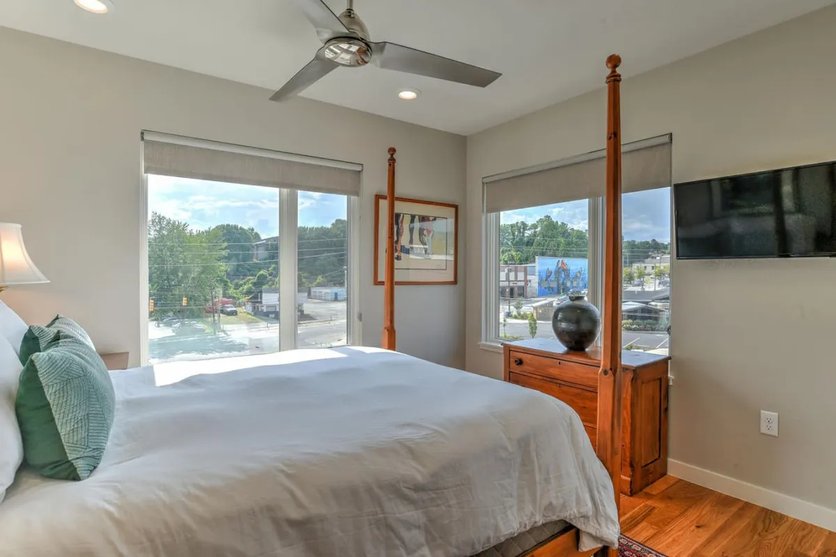 A restful upper-level room with a four-poster bed, large windows, wooden furniture, decorative artwork, and a television. The space is flexible, well-lit, and designed for both leisure and potential extra sleeping arrangements.