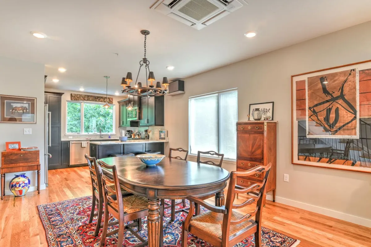 A warm dining area where a stylish table sits under an elegant chandelier, framed by striking artwork and with a clear view into the kitchen. The space invites gatherings for dinners, celebrations, or casual breakfasts.
