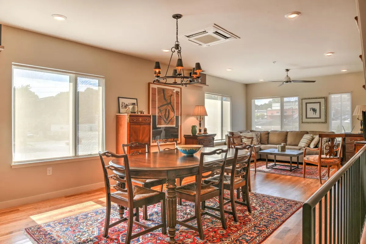 A spacious dining area with a large table set beneath a striking chandelier, surrounded by comfortable seating and contemporary artwork on the walls. Natural light pours in from nearby windows, creating a bright and welcoming space ideal for socializing and family gatherings.