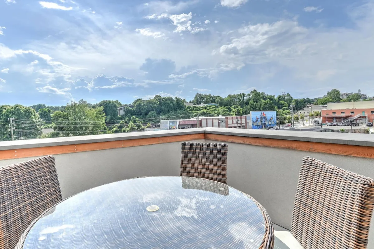 An inviting outdoor table with a glass top is surrounded by woven chairs, set on a patio with a scenic view of the surrounding landscape. The setup is ideal for relaxing with a drink or enjoying a morning coffee while soaking in the sights and sounds of the neighborhood. The overall mood is tranquil and social, with a subtle connection to the broader environment outside the property.