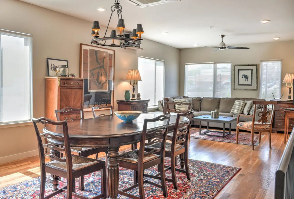 The image showcases a stylish, inviting living and dining area featuring a modern dining table beneath an elegant chandelier, a comfortable sofa for relaxing, tasteful artwork adorning the wall, and a side table that adds to the cozy ambiance. The scene exudes warmth and is perfect for gatherings, family dinners, or unwinding after a day exploring Asheville.