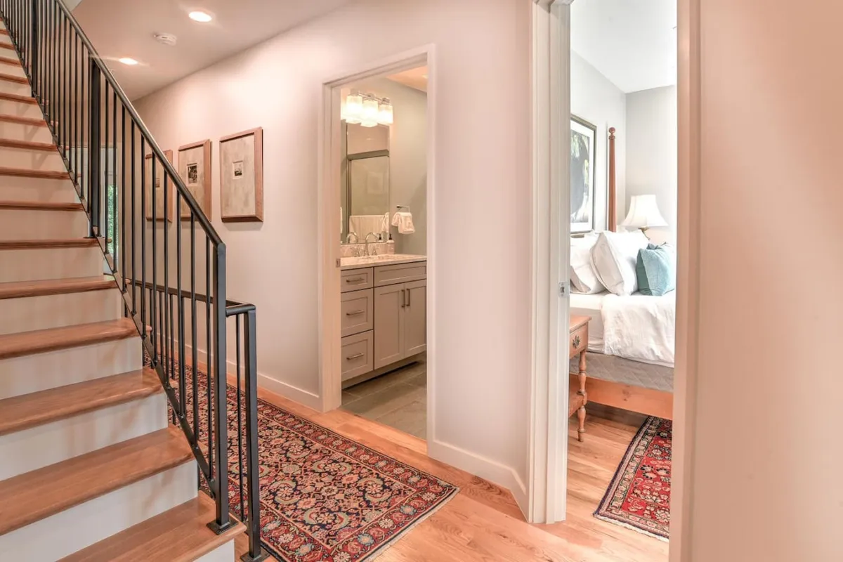 A hallway view showcasing a modern staircase leading to different levels of the home, a glimpse into a cozy bedroom, and the entrance to a bathroom. The space is accented by a decorative rug and bathed in natural light, suggesting ease of movement between areas.