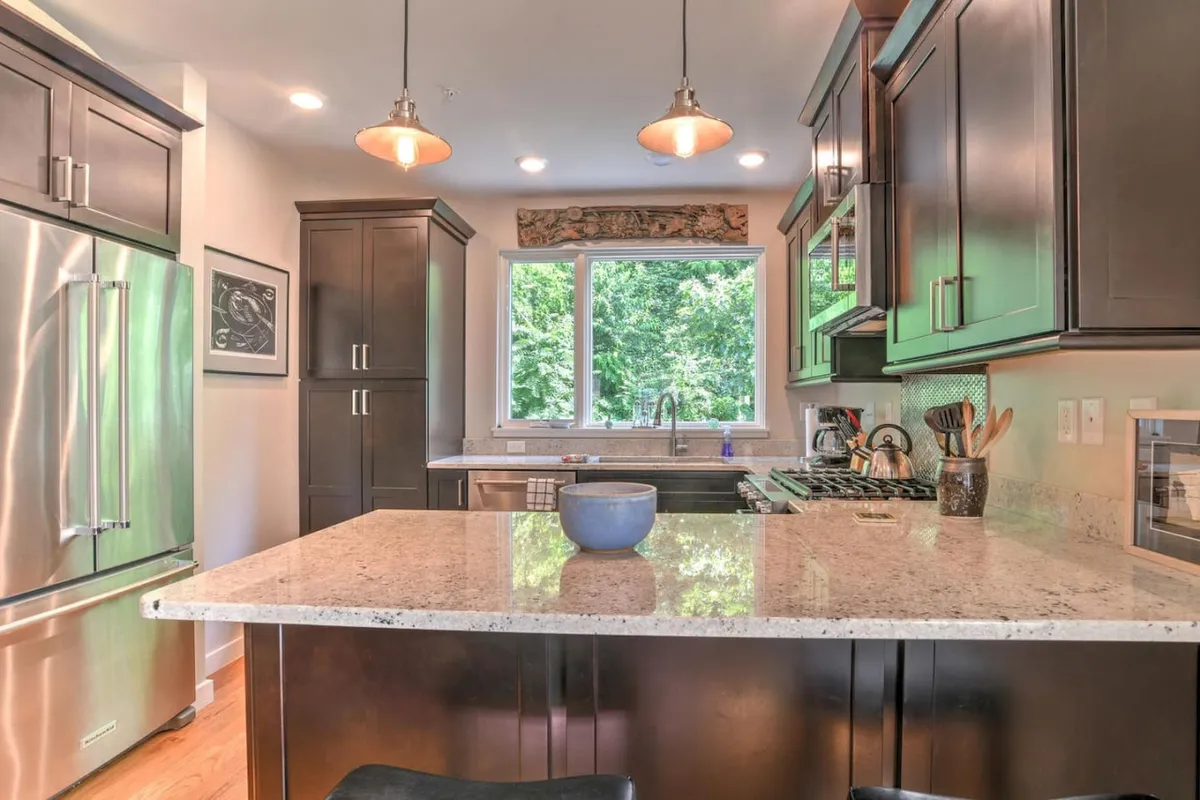 A contemporary kitchen filled with natural light from a large window, featuring granite countertops, stainless steel appliances, and sleek modern cabinetry. The space feels welcoming and lively, perfect for cooking or gathering with friends and family.