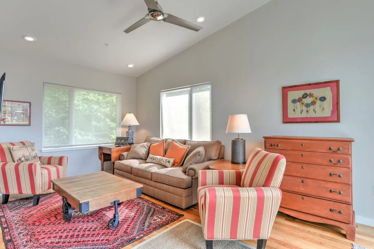 The image shows a comfortable seating area with striped armchairs, a wooden coffee table, a sofa with decorative pillows, and artwork on the wall. The large window suggests natural light and a possible view outdoors, ideal for enjoying coffee or unwinding with friends.